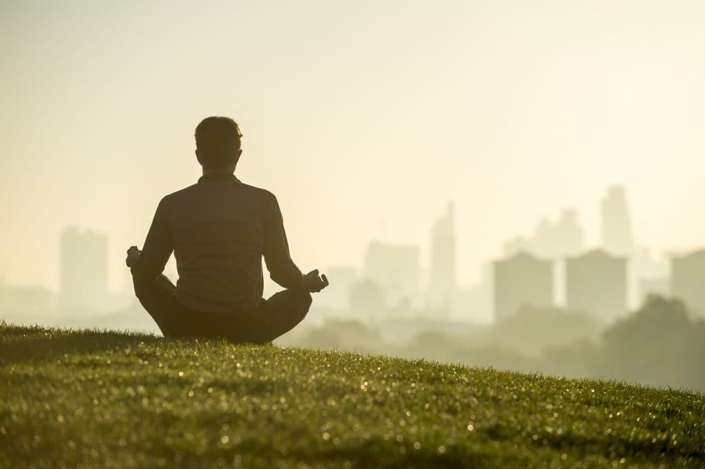 Silhouette of a man sitting in the lotus position meditating on the grassy top of Primrose Hill in front of a misty golden sunrise view of the London city skyline monday motivation setting intention